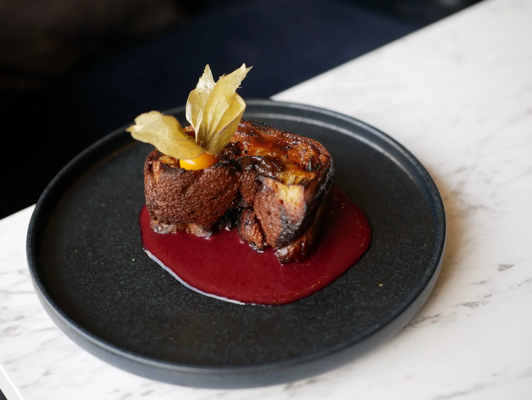 Dessert at Café Gourmand: caramelised brioche on a red berry coulis, served on a black plate.