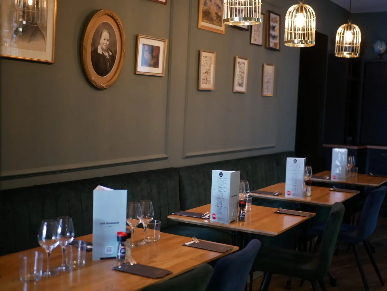 Inside Café Gourmand: green velvet banquette, set tables and vintage pictures beneath brass pendant lights.